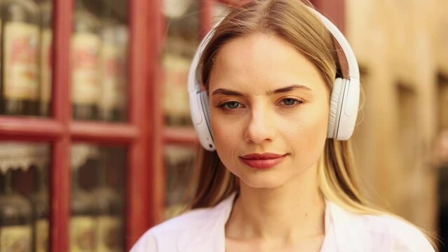 Portrait of attractive blond woman in headphones smiling while standing in the city street. Beautiful woman looking ahead outside.