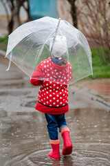 A cute little girl in a red cape, red boots and a white hat jump in puddles and has a fun.The girl has a transparent umbrella in her hands. Happy childhood. Early spring. Emotions.