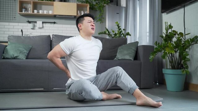 Young asian man exercise Stretch the back pain at home indoors. Athletic fitness male suffering feel hurt sudden ache shoulder and joints Muscle strain after her routine workout sitting on a yoga mat