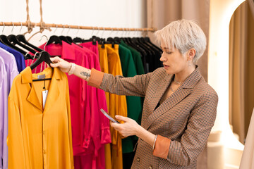 Young fashion woman buying clothes at store, standing at hangers with colorful outfit, using smartphone