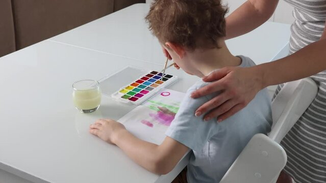 Rear view of little boy painting with paints and paintbrush with his mom. Boy learns to draw with watercolor paints together with a caring mother. Development of creative potential in young children.