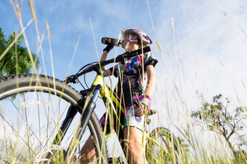 Woman drinking water from sport bottle, stands with yellow mountain bike in grass.