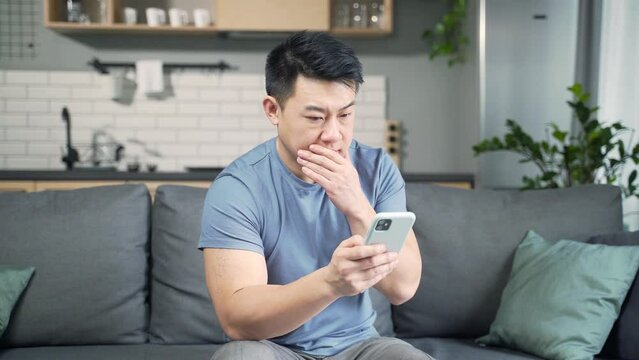 Asian man reading sms bad news on smartphone phone sitting on sofa at home. indoor. Frustrated looking at smartphone screen, feeling disappointed with received message. annoyed bank loan notification