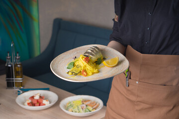The waiter holds a dish in his hand on the background of the interior of the restaurant