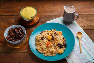 A large dish of couscous on a wooden table on a napkin with a wooden spoon next to cereals in a bowl of nuts and a jug of fruit broth.