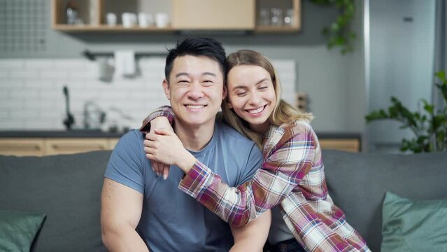 Cheerful portrait of multi ethnic married couple embracing and looking at camera while sitting on sofa at home in living room. indoor. Happy loving asian family smiling hugs. Love and affection