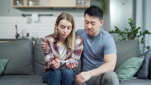 Sad upset couple after negative pregnancy test result. worried young woman and asian man holding stick while sitting on couch in home. husband comforting supports wife. Disappointed together depressed