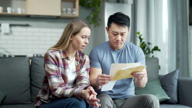 Upset Worried Multiracial couple, Asian man and woman, together at home, received a letter read with bad news, frustrated and sad sitting on the couch at home. unpaid bills and taxes feels stressed