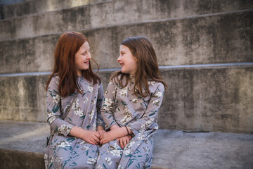 Two preteen sisters sitting in outdoor amphitheater smiling at each other; girls wear similar dresses