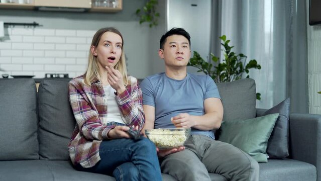 Bored mixed race asian couple at home choose a television show to watch and switch channels with a TV remote control. Young family husband and wife cannot choose a show. watching news or film