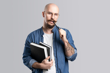 Portrait of a handsome nerd guy on a gray background, holds a books and pen in his hand with a serious face and thinks.