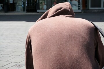 Junger Mann mit rosa Kapuzenpullover sitz von hinten auf Bank in Stadt bei Sonne im Frühling 