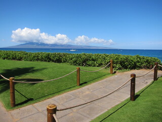 Concrete walkway towards the ocean, wooden stanchions with brown ropes, trimmed green lawn, a hedge, deep blue sea, clear blue sky. Lanai island beneath white clouds. Kaanapali, Maui, Hawaii.