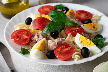 Canned tuna salad with pasta, cherry tomatoes, black olives, boiled egg and basil on gray background