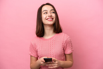 Young Ukrainian woman isolated on pink background using mobile phone and looking up