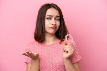 Young Ukrainian woman holding invisible braces isolated on pink background making doubts gesture while lifting the shoulders