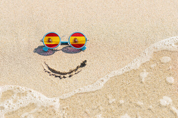 Fototapeta na wymiar A painted smile on the beach and sunglasses with the flag of Spain. The concept of a positive holiday in the resort of Spain.