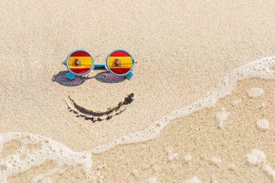 A painted smile on the beach and sunglasses with the flag of Spain. The concept of a positive holiday in the resort of Spain.