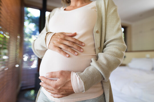 Mid section of caucasian pregnant woman touching her belly standing at home