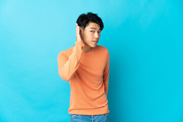 Young Chinese man isolated on blue background listening to something by putting hand on the ear