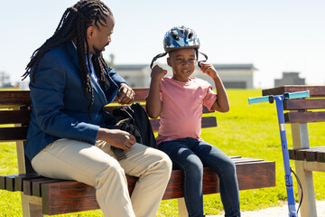 African american businessman sitting on bench with son wearing cycling helmet on sunny day