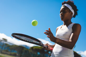 Young female african american tennis player playing with racket and ball at court against sky - Powered by Adobe