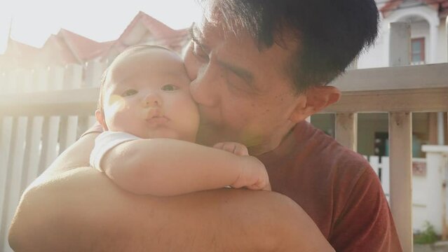 CloseUp Asian smile 60s retired old man hugs and kiss on a chubby face of newborn baby in his arms. Grandpa Comforting his nephew against summer warm light, residential village in background 