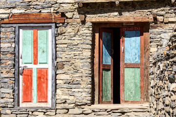Fototapeta na wymiar Multicolour red orange turquoise blue green distressed wooden shutters on stone House wall in abandoned village near Manang, Nepal. Authentic traditional buddhist architecture