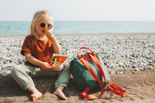 Child girl with lunchbox eating vegetables outdoor travel vacation healthy lifestyle vegan food picnic on beach hungry kid with lunch box snacks and backpack