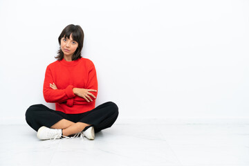 Fototapeta premium Young mixed race woman sitting on the floor isolated on white background with arms crossed and looking forward