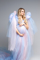 Fototapeta na wymiar A beautiful pregnant girl in a blue airy dress on a gray background. Angel. Vertical photo.