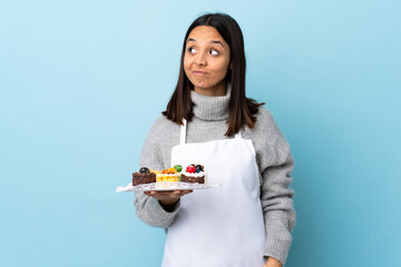 Pastry chef holding a big cake over isolated blue background having doubts while looking up.