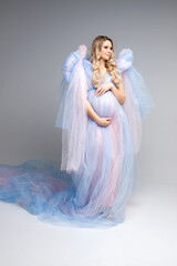 Beautiful pregnant girl in a blue dress. Pregnant girl with blonde hair on a gray background. Airy light dress. Angel. Vertical photo. Studio shooting.