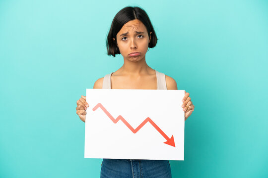 Young mixed race woman isolated on blue background holding a sign with a decreasing statistics arrow symbol with sad expression