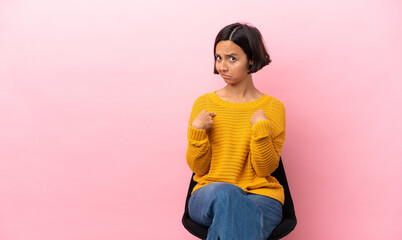 Young mixed race woman sitting on a chair isolated on pink background pointing to oneself