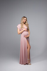 Fototapeta premium A beautiful pregnant woman holds her hands on her stomach on a gray background. A young woman in a pink maternity dress is waiting for the birth of a child. The concept of the pregnancy holiday.