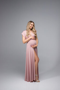 A beautiful pregnant woman holds her hands on her stomach on a gray background. A young woman in a pink maternity dress is waiting for the birth of a child. The concept of the pregnancy holiday.