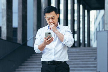Fototapeta premium Serious and pensive Asian businessman typing on the phone, a man outside the office