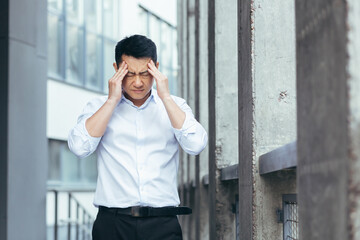 Tired and sad Asian has a severe headache, a businessman outside the office, holding hands behind...