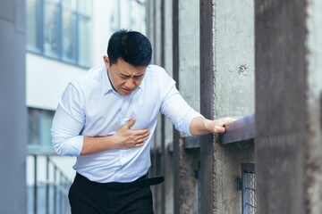 Asian office worker sick, having severe chest pain, businessman holding hands on heart outside...