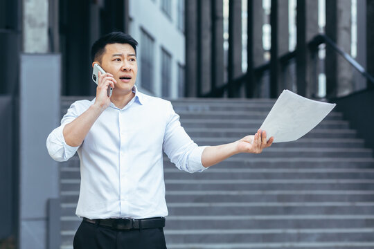 Frustrated insurance agent with documents talking on the phone outside the office, depressed Asian businessman