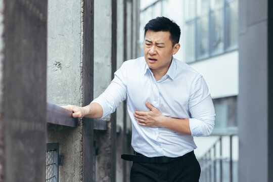 Asian office worker sick, having severe chest pain, businessman holding hands on heart outside office