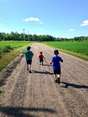 Fototapeta na wymiar Three boys run away from camera on a gravel road lined on both sides by green fields topped by blue sky summer day in rural northern Wisconsin.