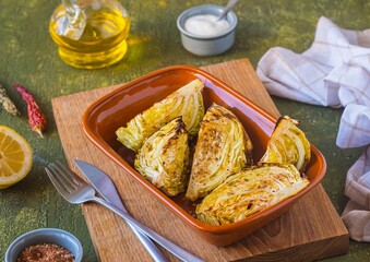Fresh young cabbage baked in pieces in the oven with spices and olive oil in a ceramic form on a green concrete background. Vegan food. Vegetable recipes