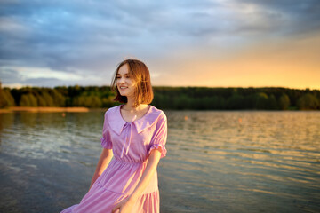 Beautiful teenage girl wearing pink dress having fun by a lake on warm and sunny summer day. Pretty young girl on a sunset.