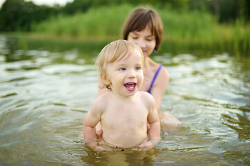 Cute toddler boy and his big teenage sister playing by a river on hot summer day. Adorable child...