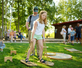 Little girl trying to walk on giant wooden feet in the park. Child playing funny outdoor game on sunny summer day. Family leisure for kids and parents.