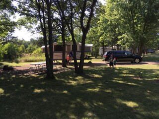 Summer camp site with three boys and an adult man in shorts and shirtsleeve shirts with a picnic table trees and a fire ring.