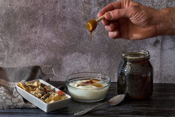 A person's hand pouring natural honey over freshly made homemade yogurt and nuts in a container