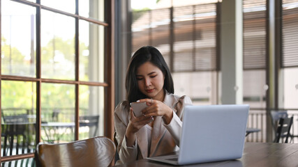 Fototapeta premium Pretty young stylish woman sitting in front of laptop at coffee shop and using mobile phone.